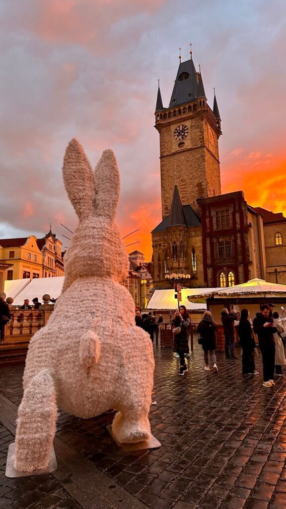 A large fluffy Easter bunny decoration stands in the foreground of Prague's Old Town Square, facing the Old Town Hall Tower. The sky is ablaze with a dramatic sunset, and the wet cobblestone streets reflect the golden light of the Easter market stalls.