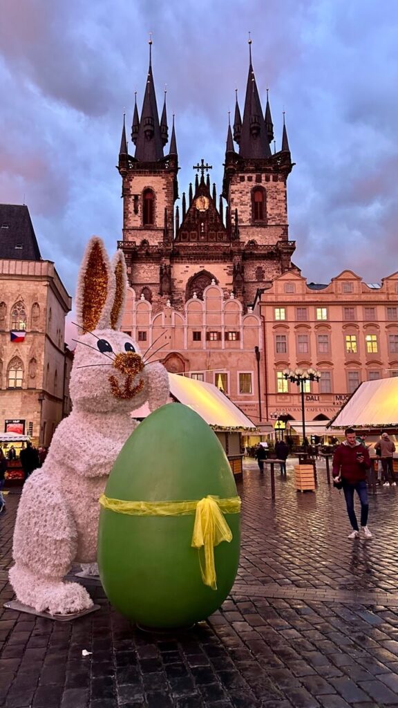 A festive Easter display featuring a large white bunny with glittery ears leaning against a giant green egg tied with a yellow ribbon. Behind it, the gothic spires of the Church of Our Lady before Týn tower over the Easter market at sunset.