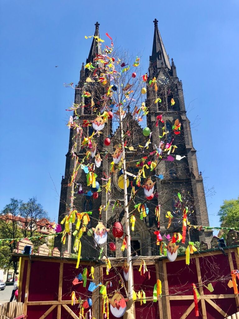 A tall Easter tree decorated with colorful ribbons, hand-painted eggs, and cheerful spring ornaments stands in front of the neo-gothic Church of St. Ludmila. Wooden stalls and festive decorations fill the square for the Easter market.