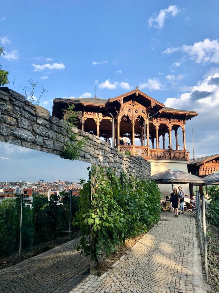 A beautifully carved wooden pavilion in Havlíčkovy Sady, standing above a stone wall with a scenic overlook of Prague. The late afternoon sunlight casts warm tones on the intricate architecture and surrounding vineyards.