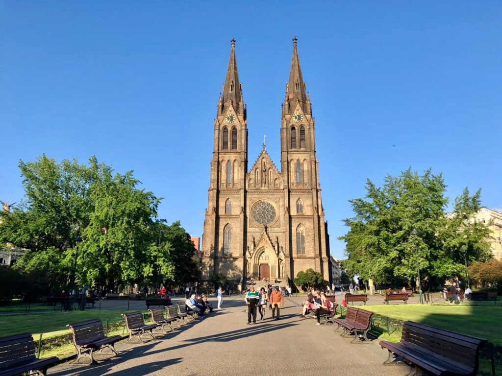 The majestic Church of St. Ludmila towers over the sunlit square of Náměstí Míru, surrounded by green trees and park benches. Locals and visitors alike enjoy the warm spring day in one of Prague’s most beautiful open spaces.