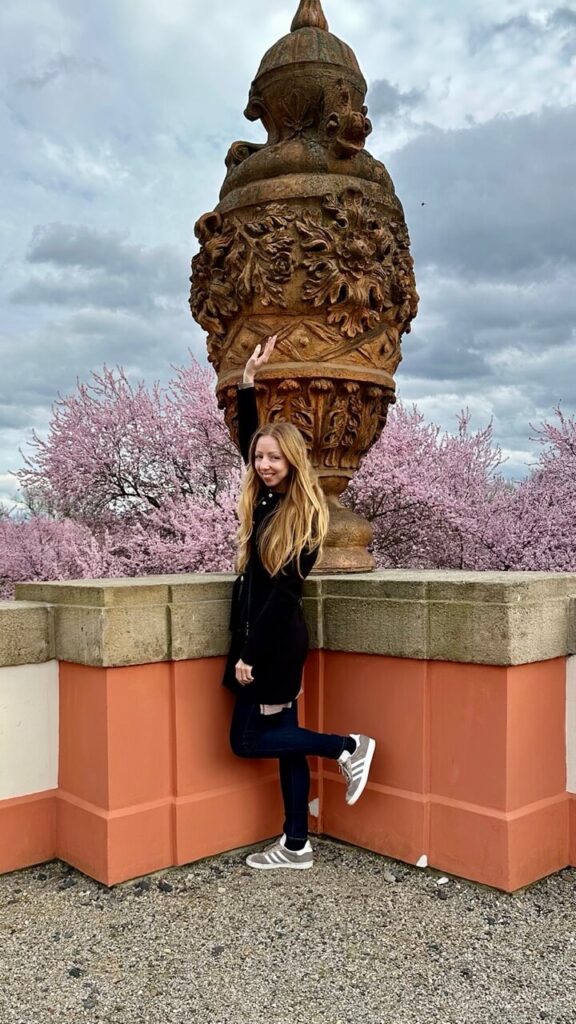 A smiling woman in a black coat playfully poses next to an ornate stone urn in the Troja Château Gardens. Behind her, cherry blossoms burst into full bloom, creating a stunning springtime backdrop.