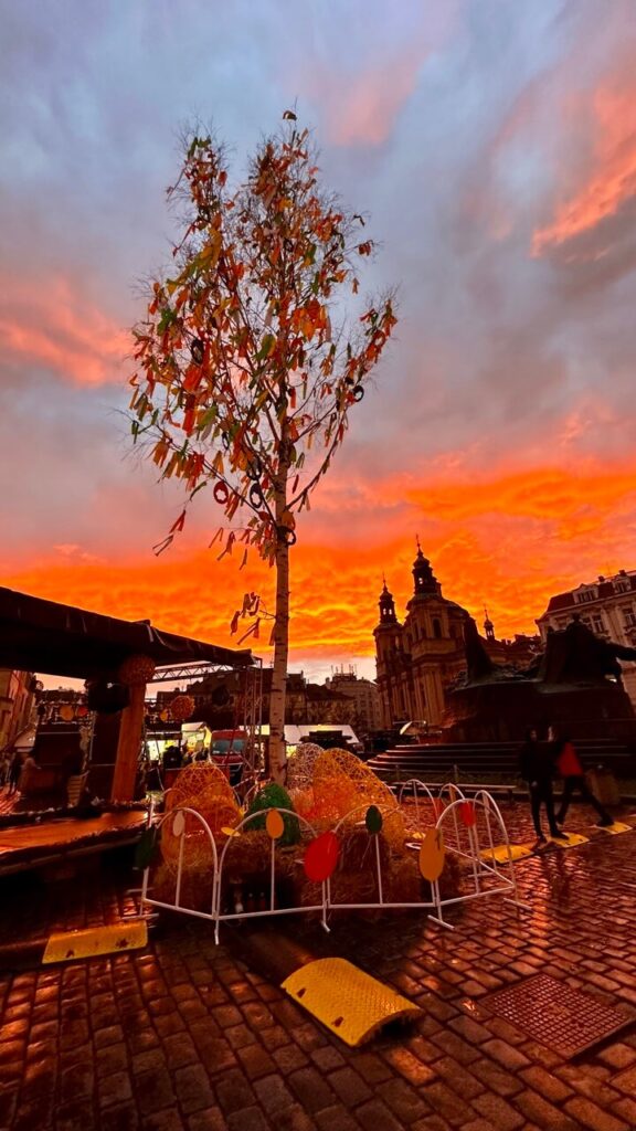 A tall Easter tree adorned with colorful ribbons and ornaments stands against a fiery orange sunset sky. The silhouette of St. Nicholas Church and the surrounding Easter market stalls create a magical atmosphere in the heart of Prague.