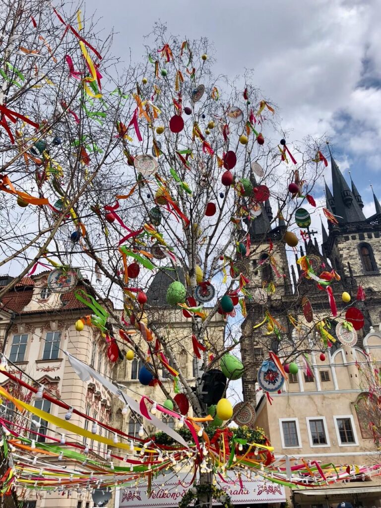 A close-up view of an Easter tree adorned with vibrantly colored ornaments, ribbons, and painted eggs. The market stalls and historic architecture of Old Town Square bring a charming springtime atmosphere.